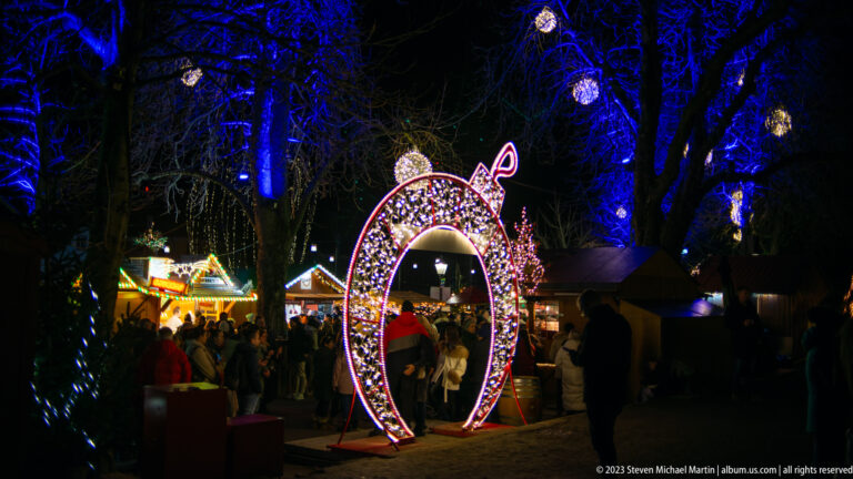 Large illuminated Christmas ornament-shaped arch with people walking through it among market stalls and blue-lit trees.