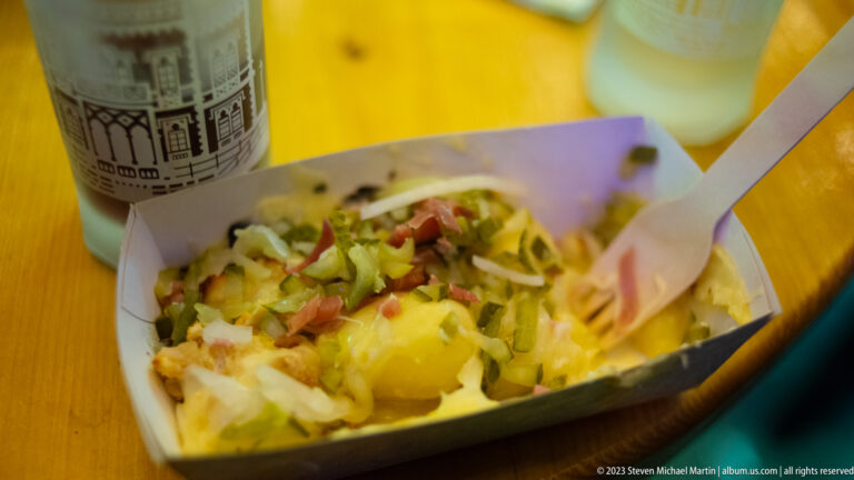 Paper tray of hot potato-based Christmas market food topped with cheese, vegetables, and meat, with a wooden fork.