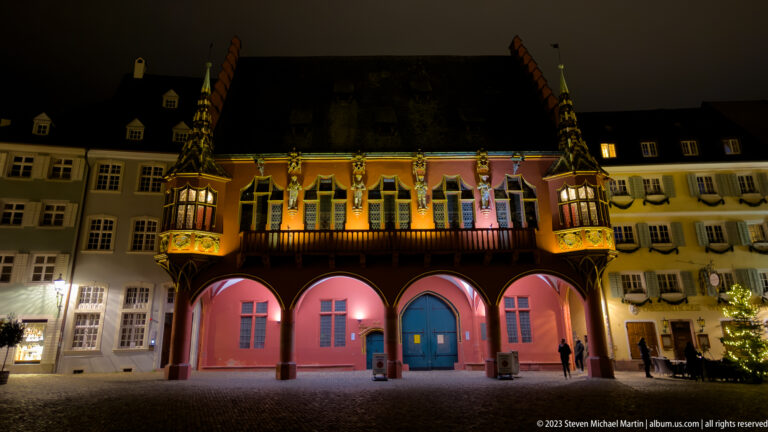 Historisches Kaufhaus at night with red facade, ground-floor arcade, Gothic windows, and projecting corner oriels.