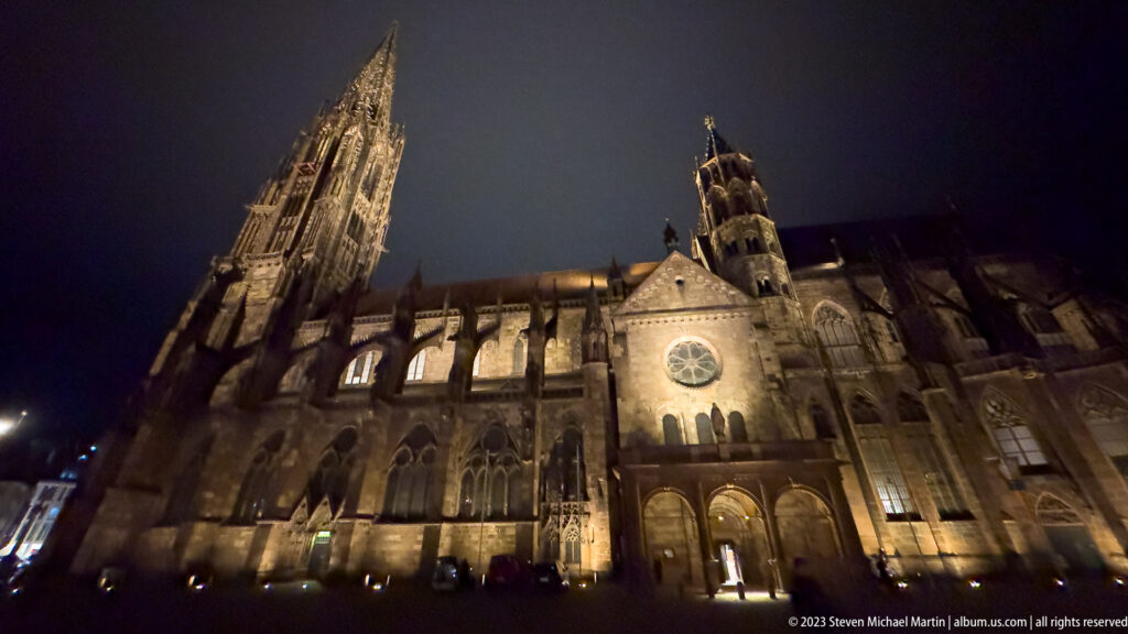 Wide-angle nighttime view of Freiburg Minster with soaring tower, illuminated Gothic nave, buttresses, and side chapels.