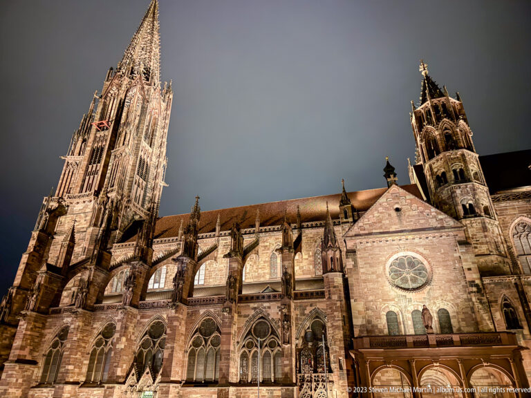 Side view of illuminated Freiburg Minster showing openwork tower, pointed windows, buttresses, roofline, and a rose window.