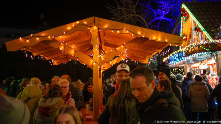 Crowded Christmas market scene beneath a wooden canopy with warm string lights and a brightly decorated stall behind.