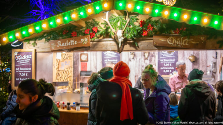 Christmas market raclette stall with green lights, evergreen garland, menu boards, customers, and staff behind the counter.