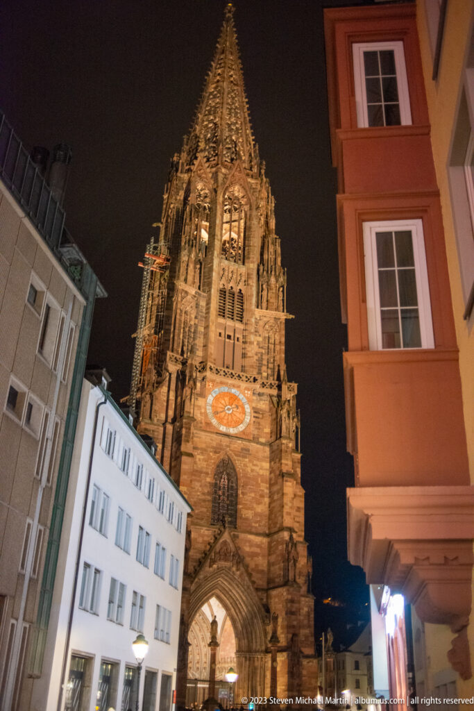 Illuminated Gothic tower of Freiburg Minster rising between Old Town buildings at night.