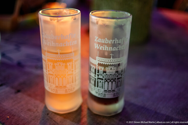 Two tall souvenir glasses printed “Zauberhafte Weihnachten,” one containing an amber drink, with Freiburg-style architectural graphics.