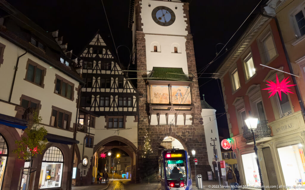 Night view of Freiburg’s Martinstor with clock tower, gate passage, tram, historic buildings, and overhead wires.