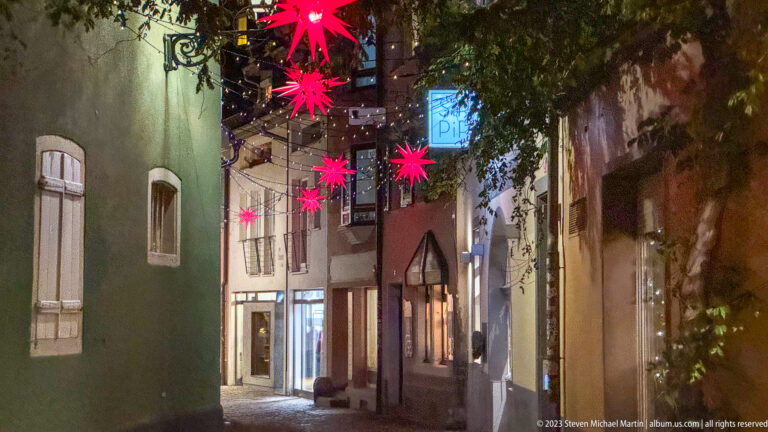 Narrow Freiburg old-town lane with red hanging star lights, colorful façades, shop windows, and overhanging foliage.