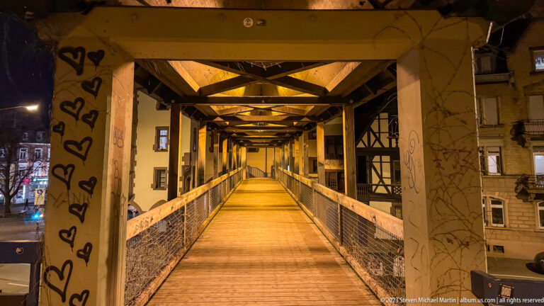 Long covered footbridge with timber trusses, metal railings, graffiti, and warm lighting at night.