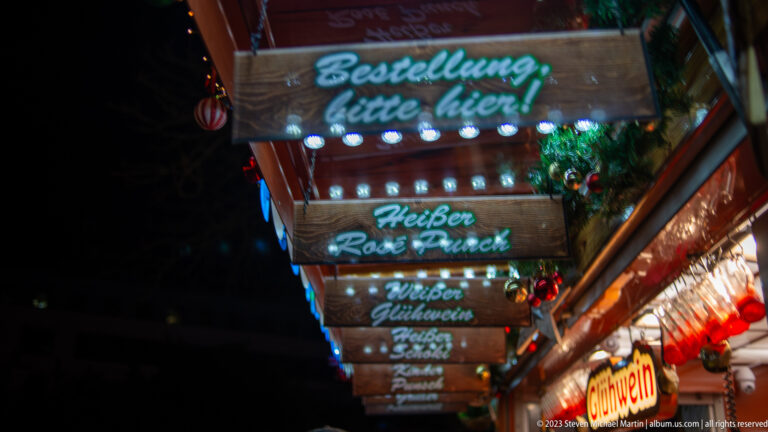 Wooden Christmas market menu boards for hot punch and mulled wine hang above a brightly lit counter.