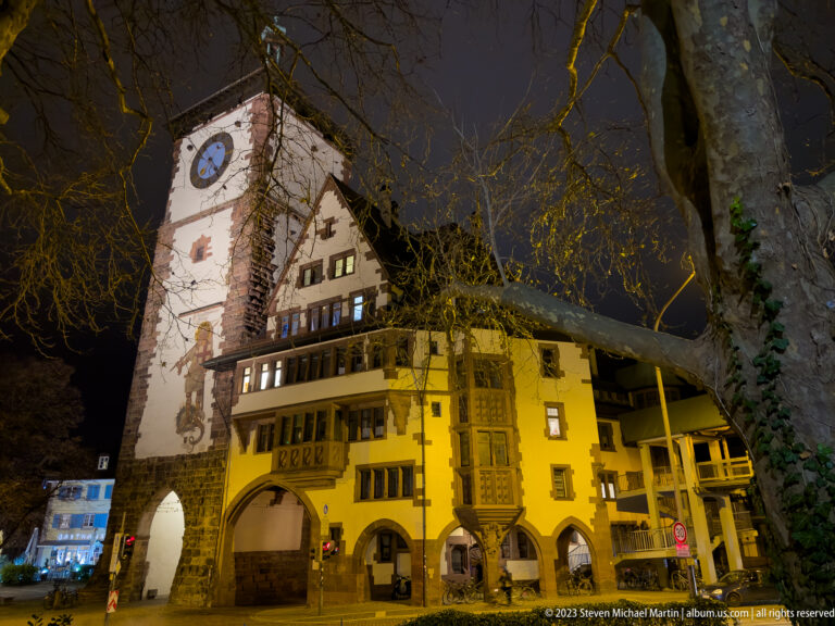 Martinstor rises behind a yellow historic building with arcades and carved bay windows on a Freiburg street at night.