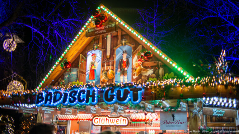 Badisch Gut Christmas market stall with multicolored lights, figures, greenery, and Glühwein signage.