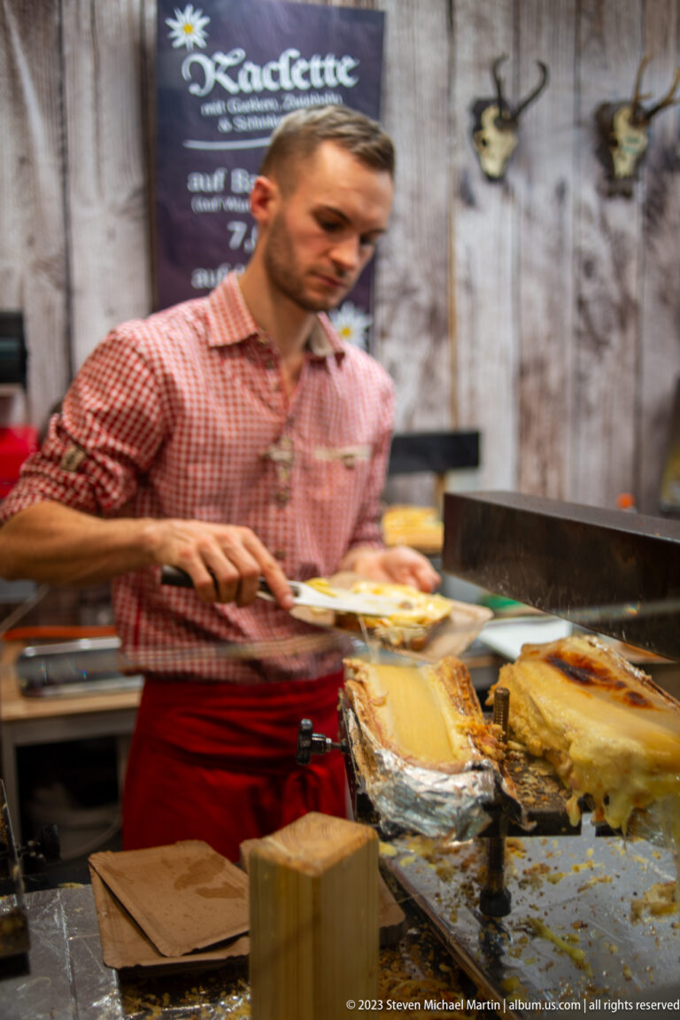 Vendor in a red checked shirt serves melted raclette cheese beside heated cheese rounds at a Christmas market stall.