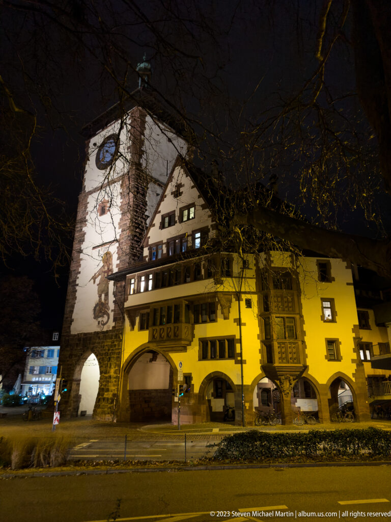 Oblique night view of Martinstor behind a yellow historic building with arcades and projecting decorative bays.