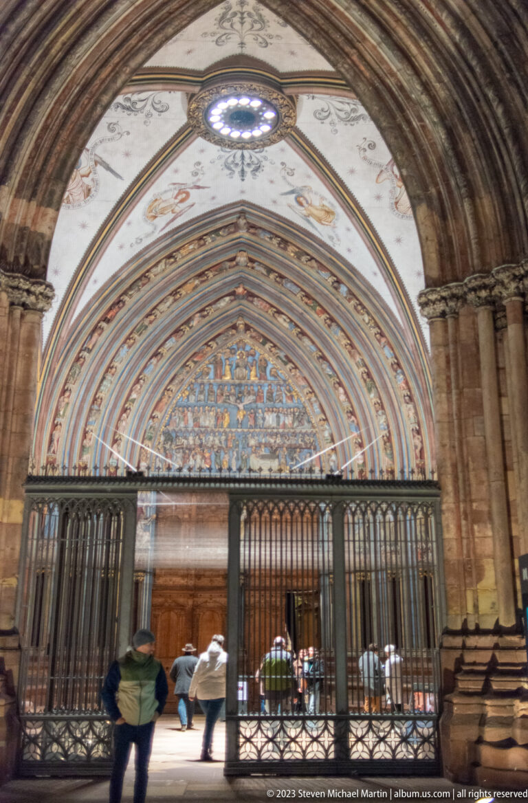 Visitors beneath Freiburg Minster’s deep Gothic west portal with painted tympanum and iron screen.