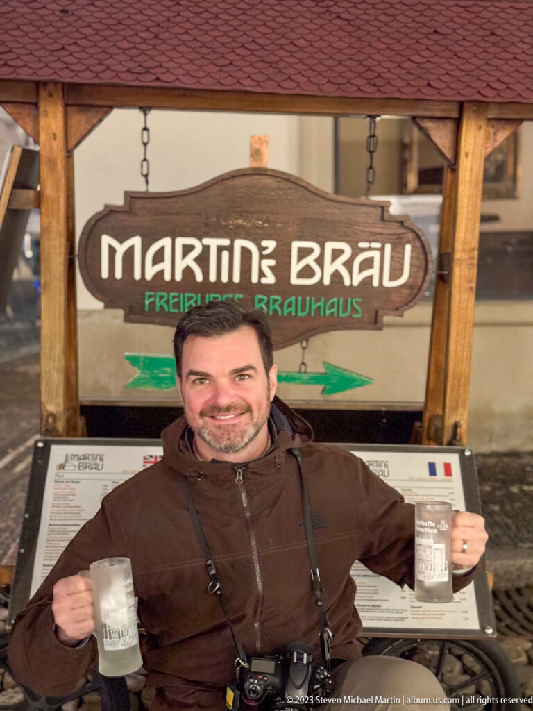 Visitor holding two beer mugs beneath the Martin’s Bräu Freiburgs Brauhaus sign, with a camera and menus visible.