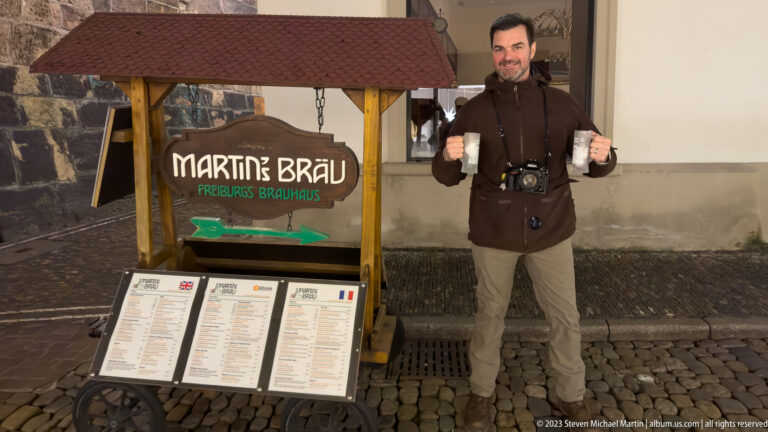 Full-length visitor holding two beer mugs beside the Martin’s Bräu sign and menu boards on a cobbled street.