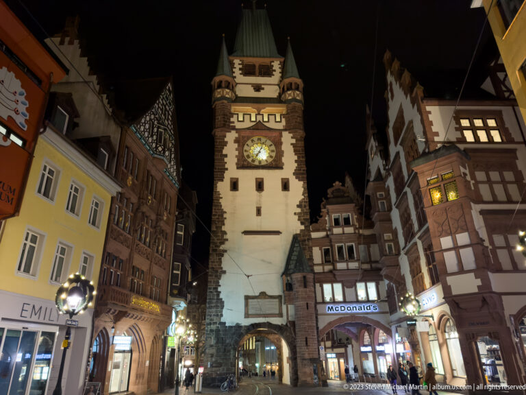 Frontal night view of Martinstor centered between historic shopfronts, with pedestrians, lights, and overhead transit wires.