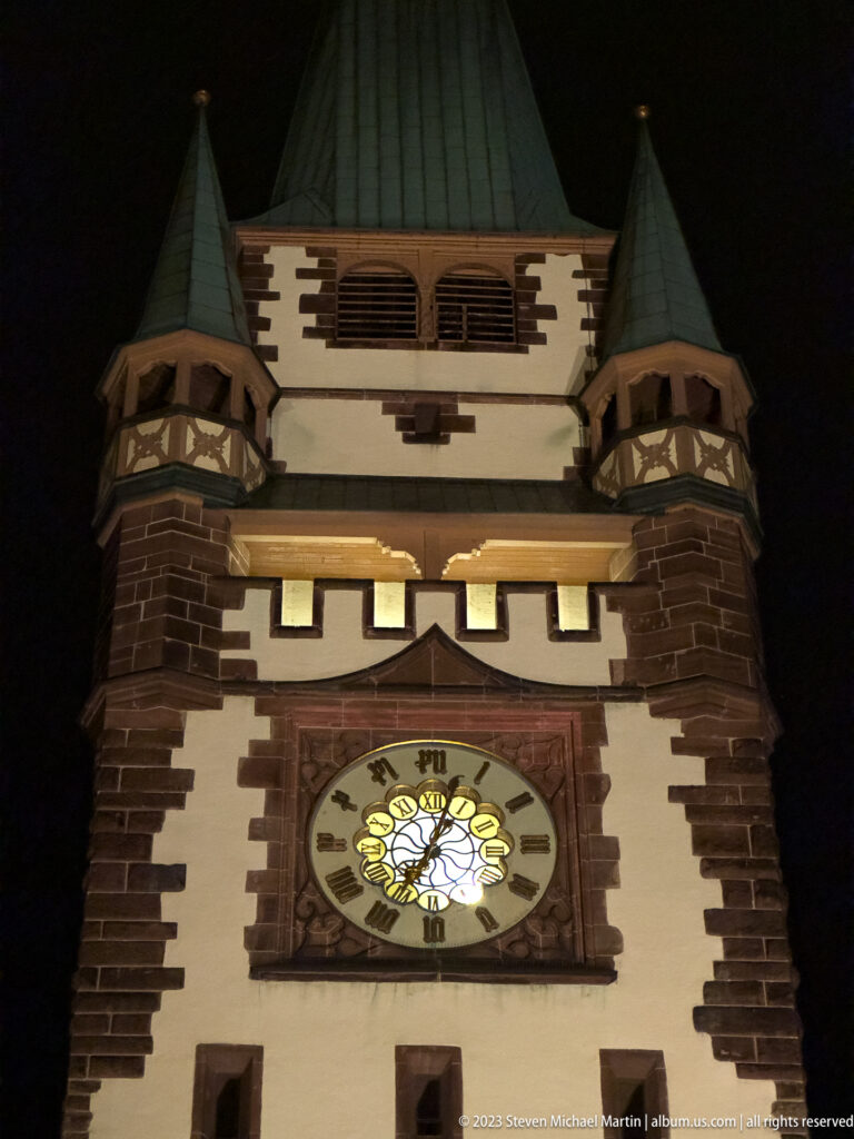 Close night view of Martinstor’s illuminated clock, white upper walls, dark trim, and steep roof with corner turrets.