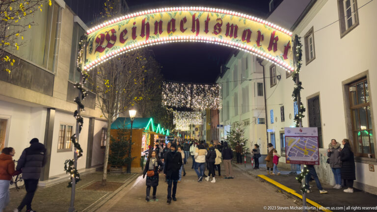 Illuminated Weihnachtsmarkt entrance arch over a pedestrian street filled with visitors, lights, and market stalls.