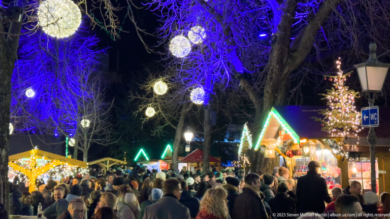 Large Christmas market crowd beneath blue-lit trees, glowing hanging spheres, timber stalls, and a decorated Christmas tree.