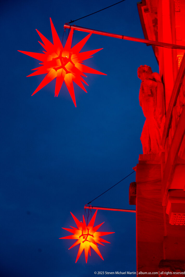 Two glowing red multi-pointed stars hang over the street beside a red-lit stone building at dusk.