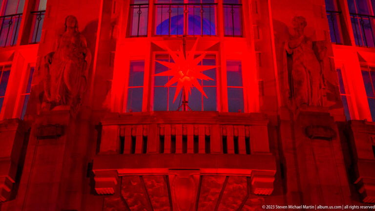 Red-lit Theater Freiburg facade with balcony, tall windows, sculpted figures, and a glowing Christmas star.