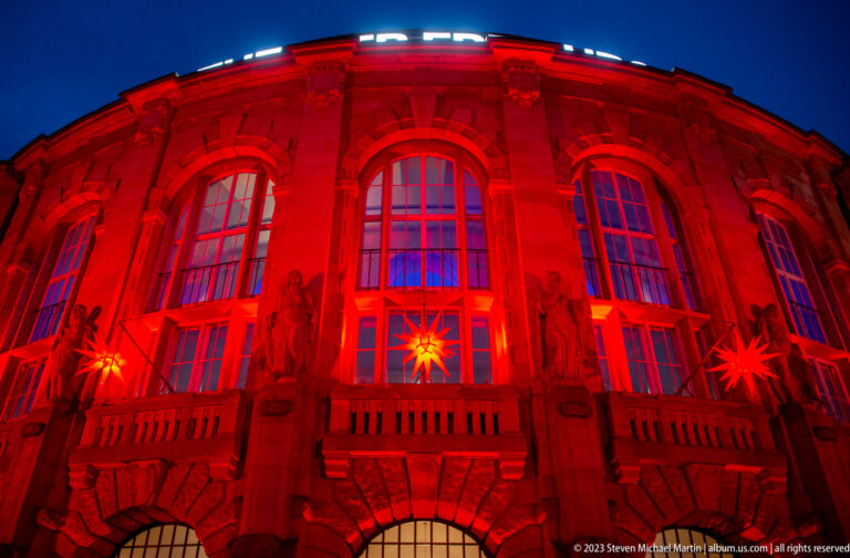 Upper facade of Theater Freiburg lit red, with tall arched windows, sculpted figures, and hanging Christmas stars.