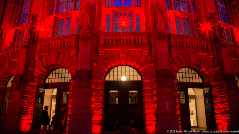 Three arched entrances of Theater Freiburg beneath a stone balcony, all illuminated red with a hanging star above.