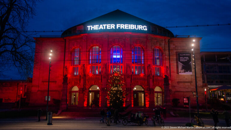 Full Theater Freiburg facade lit red at dusk, with white theater sign, arched windows, entrances, Christmas tree, and bicycles.