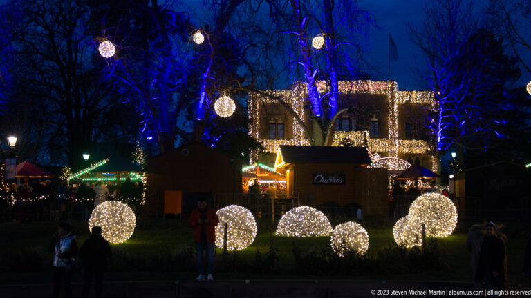 Glowing white light spheres beneath blue-lit trees, with Christmas market stalls and an illuminated building behind.
