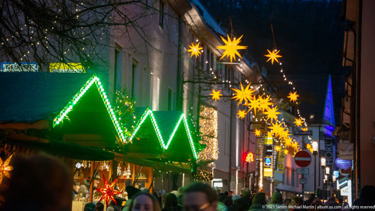 Narrow Freiburg Christmas market street with green-lit stall roofs, crowds, golden hanging stars, and a distant blue-lit spire.