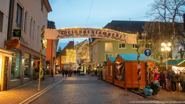 Weihnachtsmarkt entrance arch over a broad Freiburg street with timber stalls, visitors, and golden light curtains at twilight.