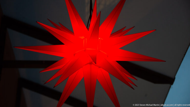Close-up of a glowing red multi-pointed Christmas star suspended beneath a dark overhang at dusk.