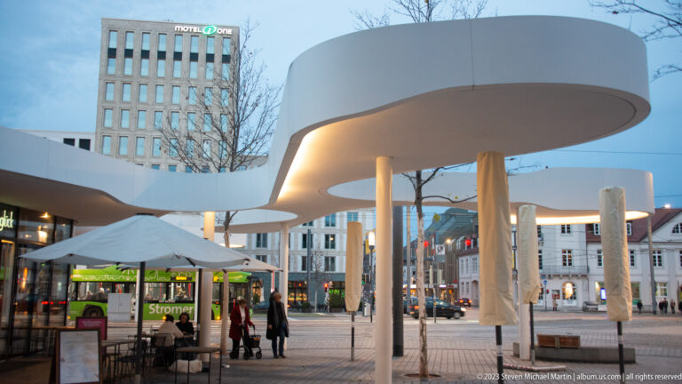 Curving white modern canopy on slender columns above a public square with café seating and pedestrians in Freiburg.