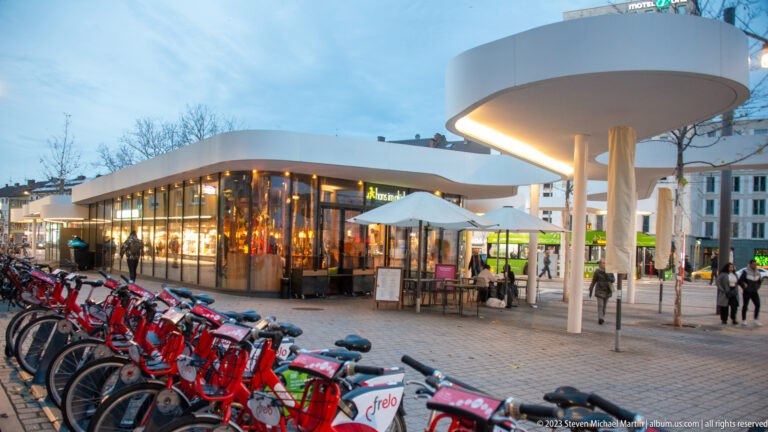 Row of Frelo rental bicycles beside a modern white pavilion, glass cafe, outdoor tables, and pedestrians in Freiburg.
