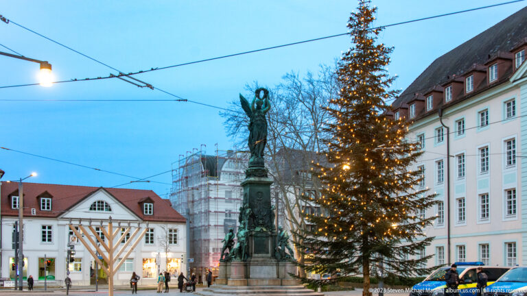 Bronze public monument beside a tall illuminated Christmas tree in a Freiburg square with historic buildings and tram wires.