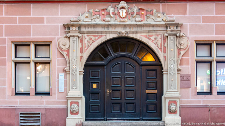 Black double door framed by an ornate pale stone portal with scrollwork and heraldic decoration on a pink façade.