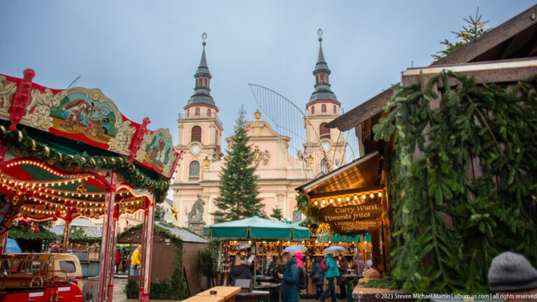 Christmas market stalls, evergreen decorations, and a tree in front of a twin-towered church in Ludwigsburg.