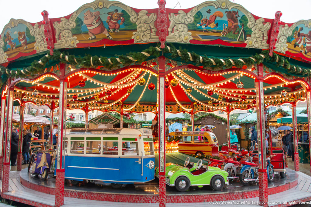 Red, green, and gold children’s carousel with rows of lights and small vehicle-shaped seats at a Christmas market.