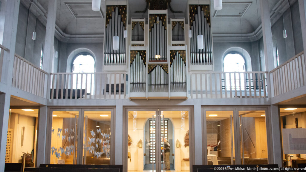 Pipe organ on a pale gallery above glass-fronted rooms in a bright white church interior.