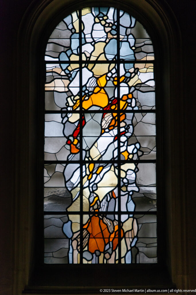 Arched stained-glass window with an abstract orange-and-yellow human figure against blue, gray, and clear glass.