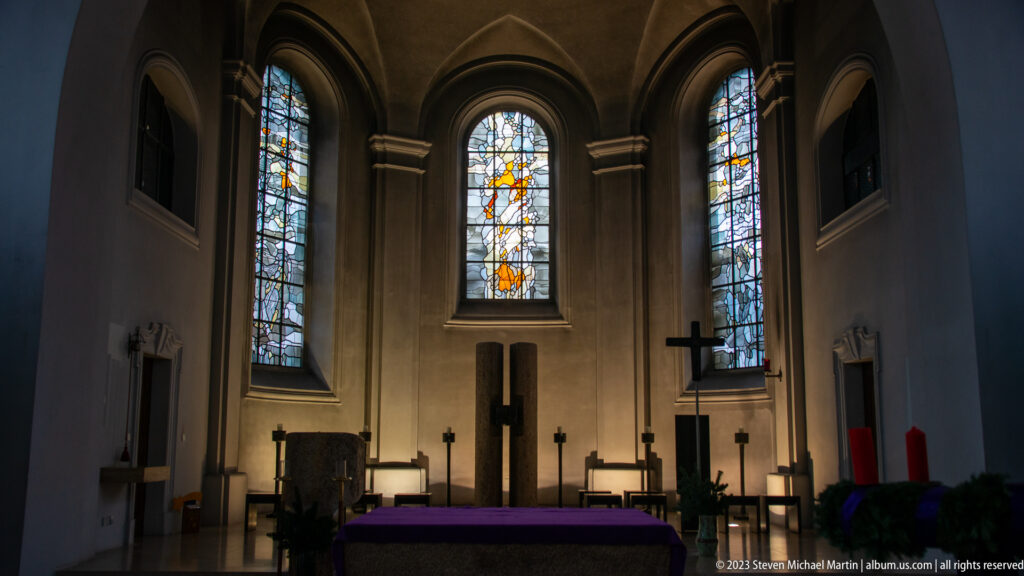Church chancel with three tall stained-glass windows, dark furnishings, a cross, and a purple-covered altar.
