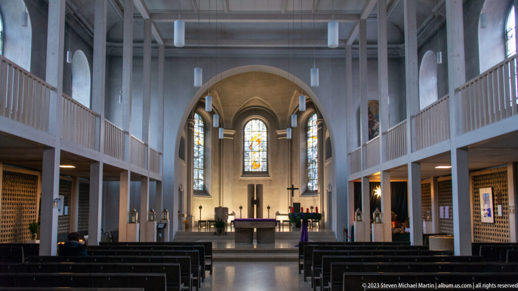 Centered view down a church nave with dark pews, white side galleries, hanging lights, and stained glass behind the altar.