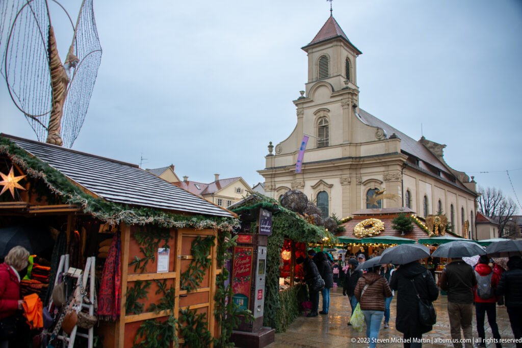 Rainy Christmas market with garland-covered wooden stalls, umbrella-carrying visitors, and a pale single-towered church beyond.