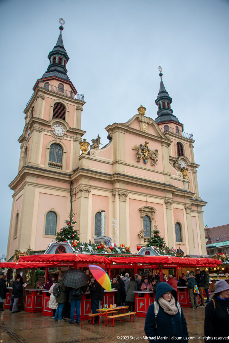 Pink baroque twin-towered church rising above red Christmas market stalls and umbrella-carrying visitors.