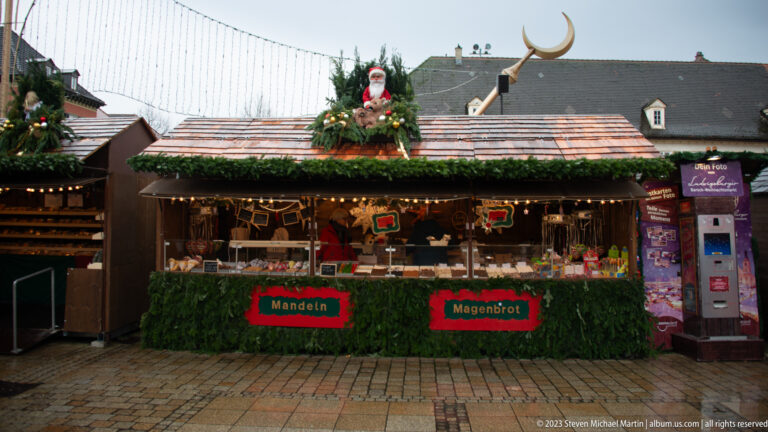 Garland-trimmed market stall labeled Mandeln and Magenbrot, with sweets displayed under warm lights and holiday decorations.