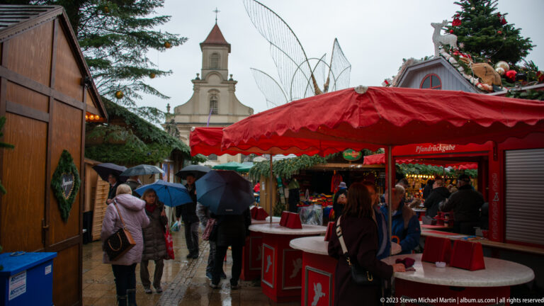 Red-canopied Christmas market stall beside a rainy lane of umbrellas, with a church tower and decorated tree in the background.