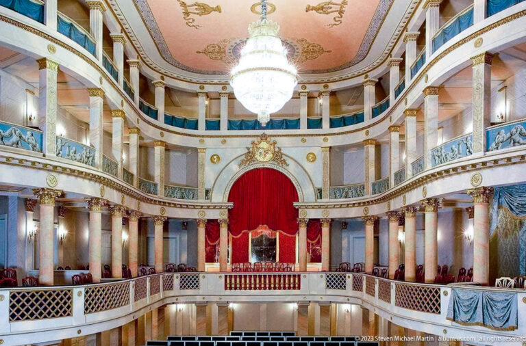 Ornate palace theater with curved galleries, red stage curtain, chandelier, gilding, and painted decorative panels.