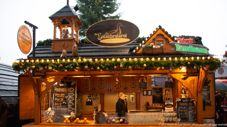Wooden Christmas market stall marked Entenbraterei, trimmed with evergreen garland, lights, carved panels, and menu boards.