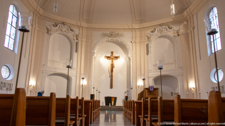 Church nave with wooden pews leading toward a large crucifix on a pale ornamented sanctuary wall.